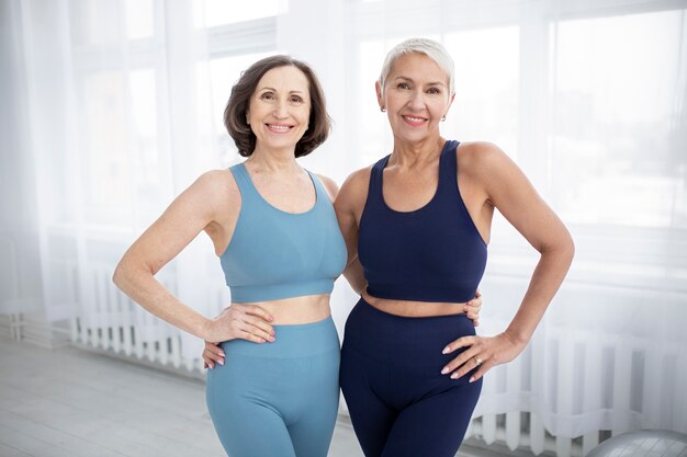 Two senior women in activewear smiling and posing indoors near window.