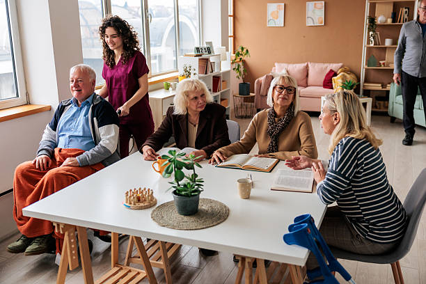 Older adults socializing at a table while a caregiver assists a seated resident in a welcoming senior living community.