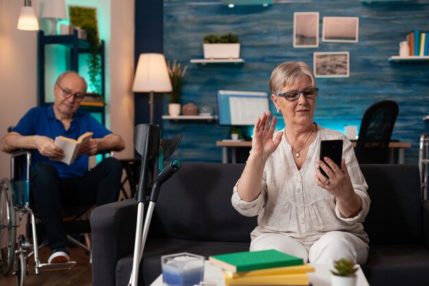 A senior woman is sitting on a sofa using a smartphone while an older man in a wheelchair reads a book in the background.